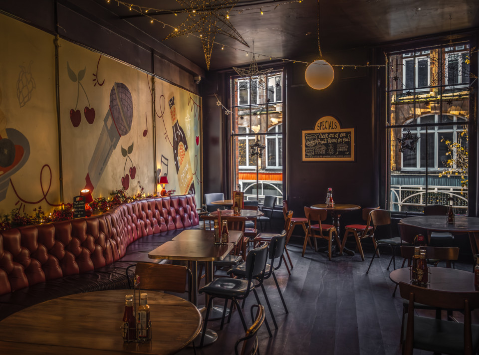 A room with assorted tables and chairs; a long wavy sofa seat is on the left and fairy lights hang from the ceiling.